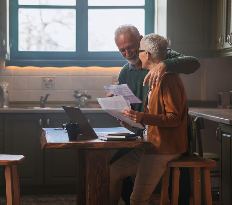 Senior couple sitting at a kitchen table, smiling and reviewing papers together with a laptop nearby.